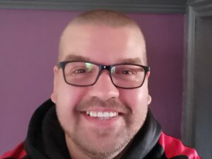 Andy smiles at the camera wearing a black hoodie and glasses. Behind him is a purple wall.