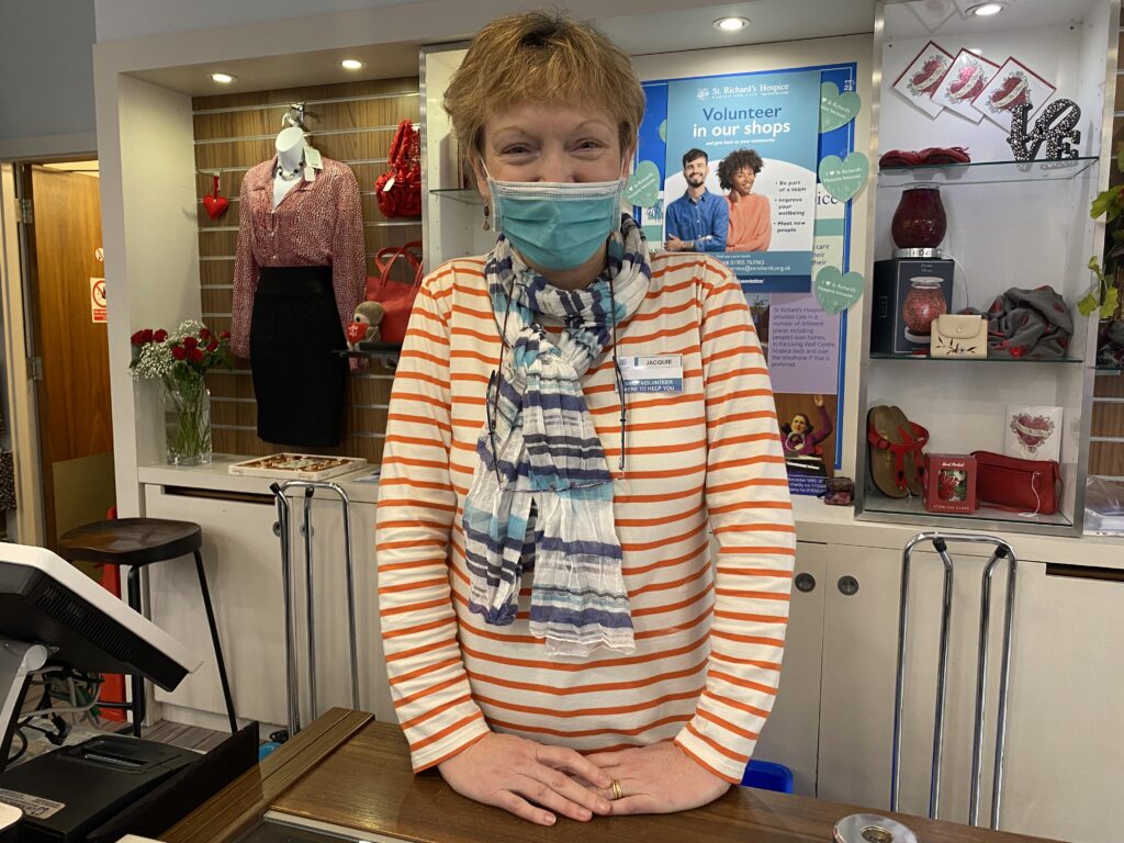 A volunteer wearing a red and white stripy top and blue face mask stands behind a shop counter. 