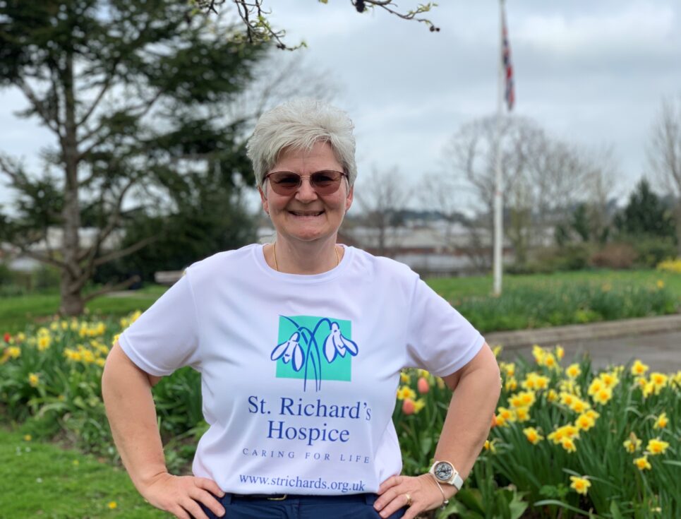 Angela McCulley stands with her hands on her hips, smiling. She is wearing a white, St Richard's Hospice branded t-shirt. Behind her are daffodils.