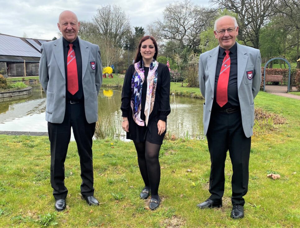 Les Janes and Art Shrimpton of Worcester Male Voice Choir stand with Kelly Johns of St Richard's Hospice in the gardens at the hospice. Les and Art wear the choir's uniform of black trousers and shirts, and grey blazers with a red tie.