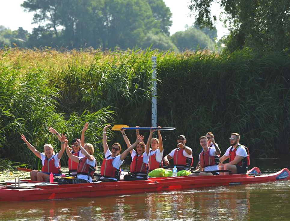 A group of people sit in two canoes on the river Avon. They are cheering, and waving their oars above their heads.