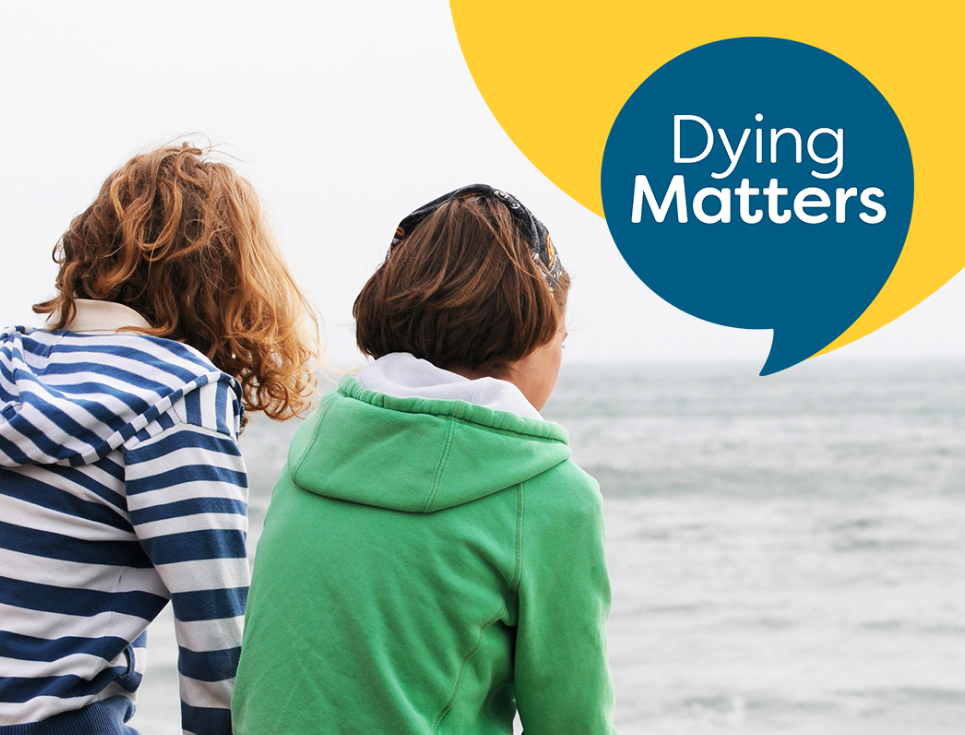 Two people sit together looking out on a grey sea. The blue Dying Matters logo is in the top right hand corner, featuring the words Dying Matters.