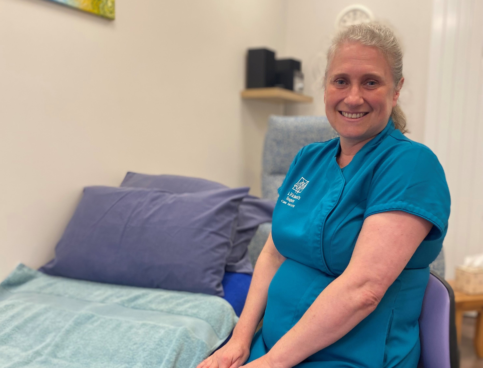 Complementary Therapist wearing a blue short sleeved top with hospice logo. Sitting next to a therapy couch