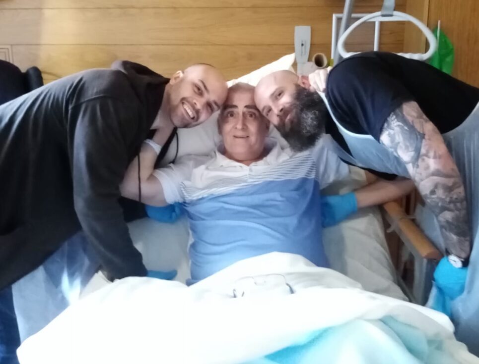 Two brothers stand either side of their dad, who is sitting in bed at St Richard's Hospice. They are all smiling.