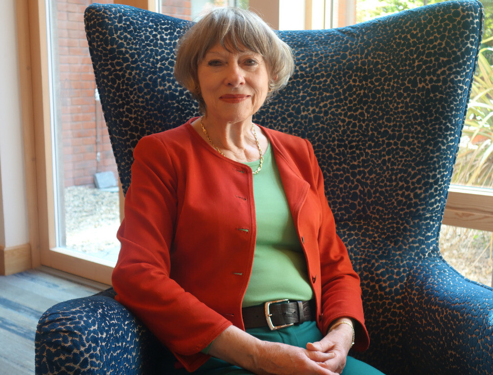 Jenny Cowpe sits in a winged armchair in the hospice Reception. She is wearing a red jacket, green top and dark green trousers.