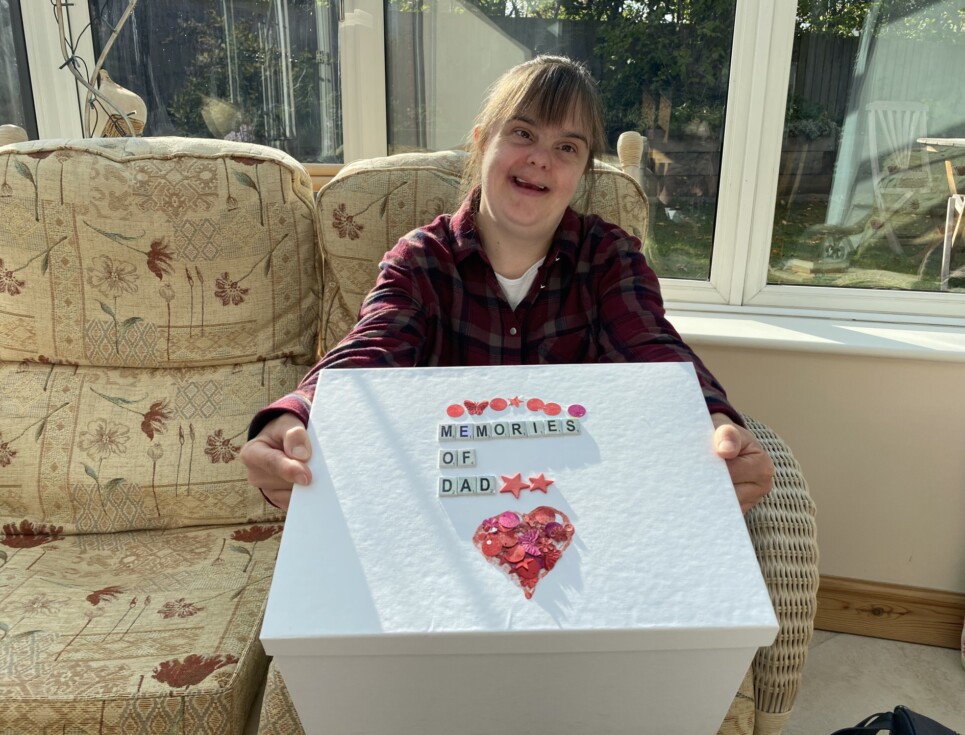 Jen sits on a sofa in her conservatory smiling and holding a white memory box decorated with the words memories of dad.