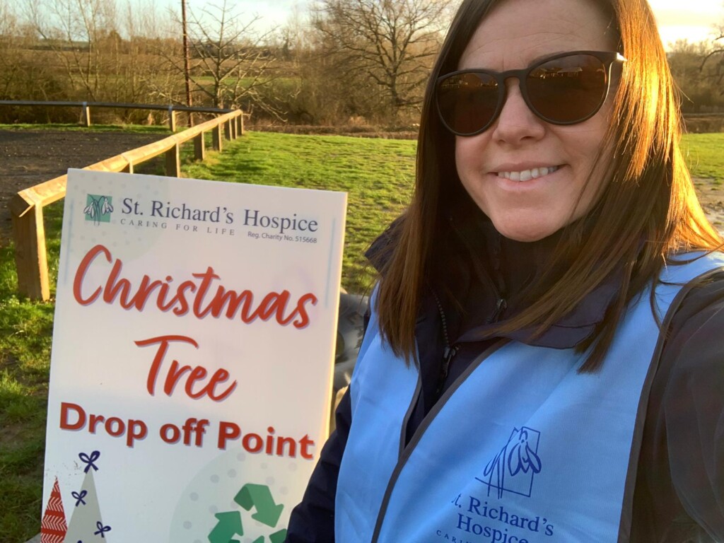 Sara stands in front of a sign which reads Christmas Tree Drop Off Point