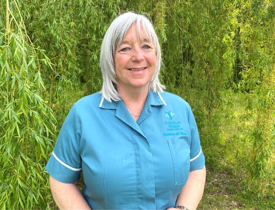 Jackie stands by a vivid green willow tree in the gardens at St Richard's Hospice. She is wearing her bright blue Hospice at Home Healthcare Assistant tunic and is smiling.
