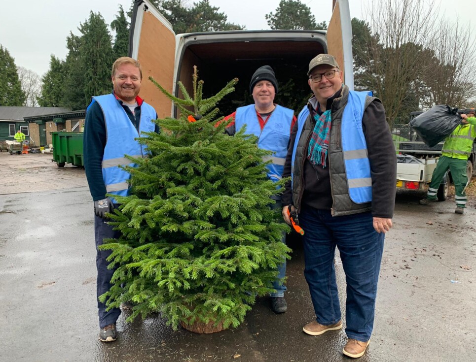 A group of three volunteers standing with a green Christmas tree in front of a van filled with trees to be recycled.