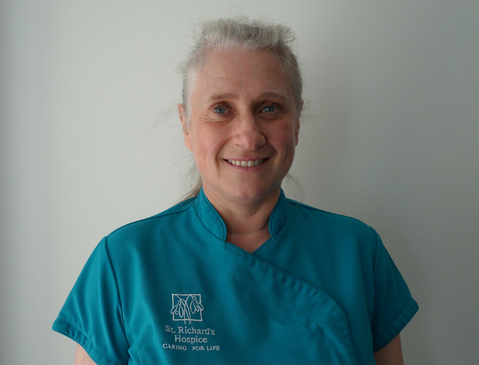 A hospice complementary therapist with her hair tied back is pictured wearing a teal uniform. She is smiling.