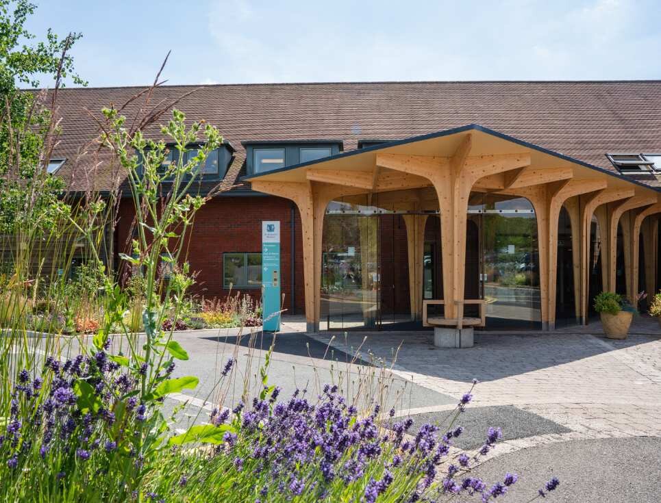 The entrance walkway into St Richard's Hospice pictured on a bright, sunny day. The walkway is constructed of wooden posts, with large sheets of glass in between.
