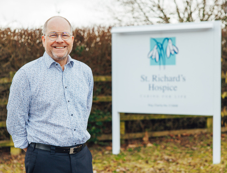 Mike Wilkerson stands by the St Richard's Hospice sign smiling.