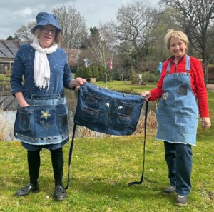 Two people wearing denim aprons over their clothes stand together in the gardens of St Richard's hospice holding a handmade, denim half apron for use while gardening.