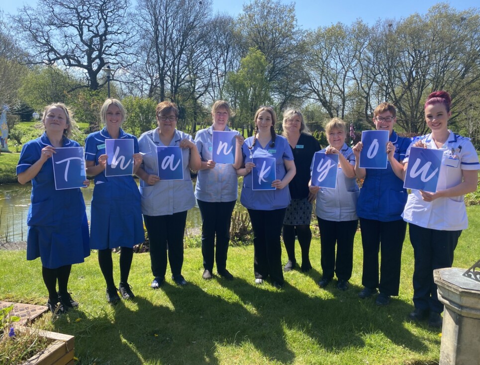 A group of nurses in blue uniforms stand together in the gardens of St Richard's Hospice on a bright, sunny day. They each hold an A4 sheet of paper with a letter on each spelling out the words thank you.
