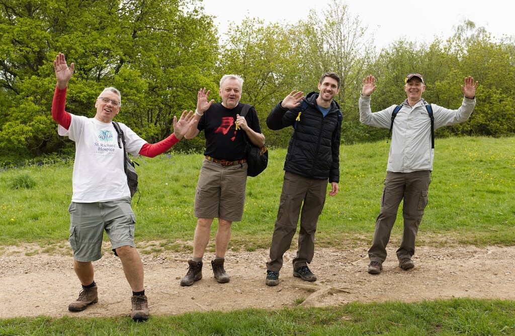 Four people taking part in the Malvern Hills Walk stop and pose for a photograph. Everyone waves at the camera.