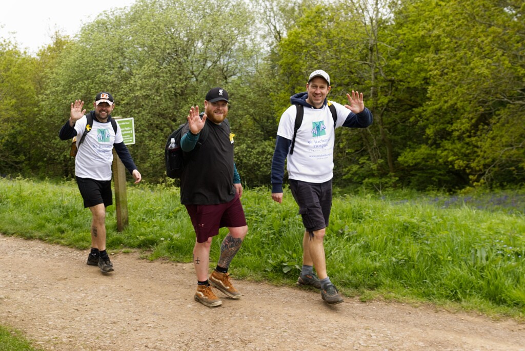 Three people taking part in the Malvern Hills Walk wave at the camera. Two are wearing white St Richard's Hospice branded t-shirts