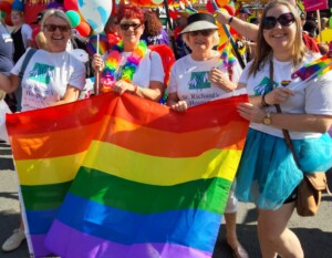 Helen, Liz, Marilyn, and Claire take part in the parade at Worcestershire Pride. Between them, they hold up a large Pride flag in rainbow colours. They are all wearing St Richard's branded t-shirts and sunglasses. They're all smiling.