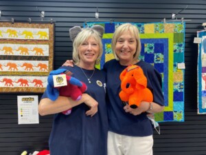 Two people, Lynn and Fiona, stand together with their arms around each other in the Worcester's Big Parade pop-up shop. They are both cuddling soft toys and Lynn is wearing toy elephant ears.