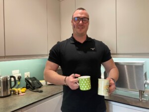 Simon, wearing a black t-shirt, stands holding two cups of tea in the bistro kitchen of the hospice's In-patient Unit. He is smiling.