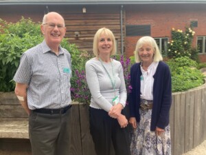 Steve, Rosalie and Heather stand together in the courtyard garden by the hospice's Living Well Centre. Flowers bloom behind them.