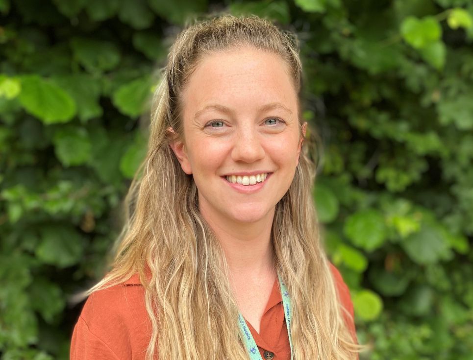 Beth pictured in front of a dark green leafy bush. She has long blonde hair is wearing an orange top. She is smiling.