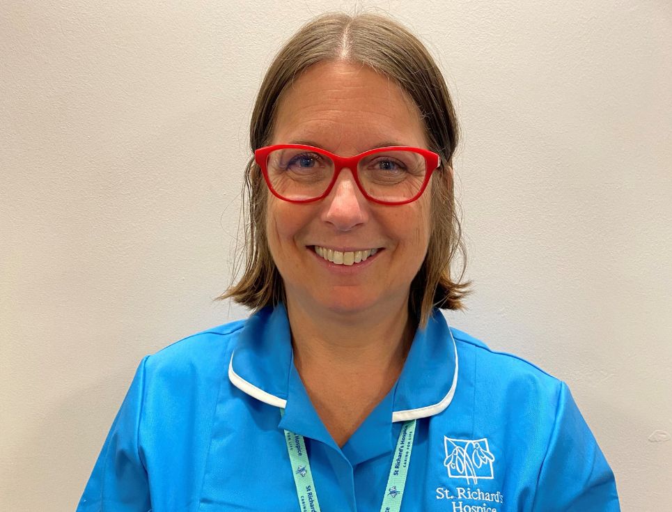 Elaine Turner, pictured against a plain white wall. She is wearing a sky blue nurses' uniform with white edging around the collar. Elaine is smiling and wears red framed glasses.