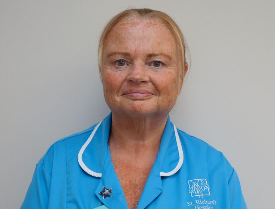 Kathy pictured against a plain, white wall. She is wearing a sky blue nurses' uniform with white edging around the collar. Kathy is smiling.