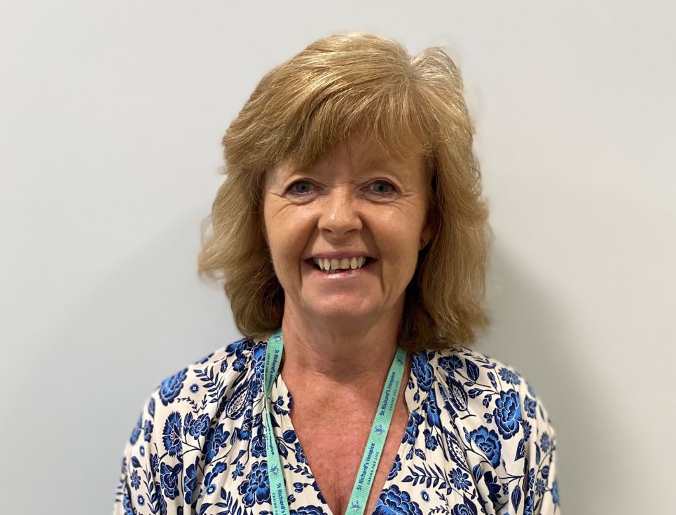 Lynn pictured against a plain white wall. She is wearing a blue and white floral patterned top and smiling.