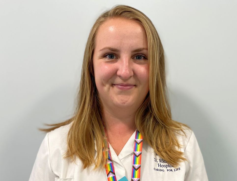 Samantha pictured against a plain white wall. She is wearing a white therapy support assistant's uniform and a lanyard featuring the multi-coloured progress Pride flag. She has blonde hair and is smiling.