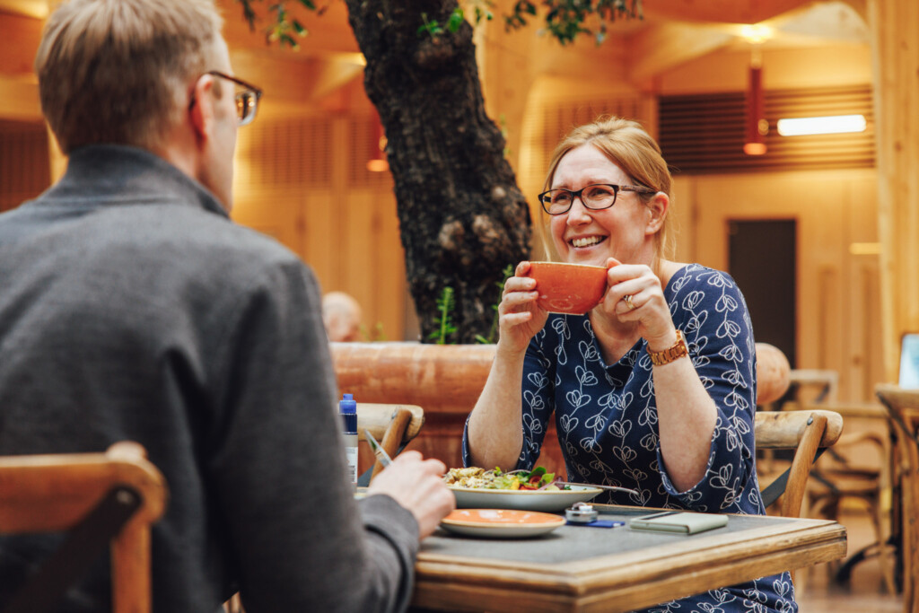 A woman sits at a table in The Green social and cafe area of St Richard's Hospice. She is holding an orange coffee cup and smiling at the person sat opposite her.