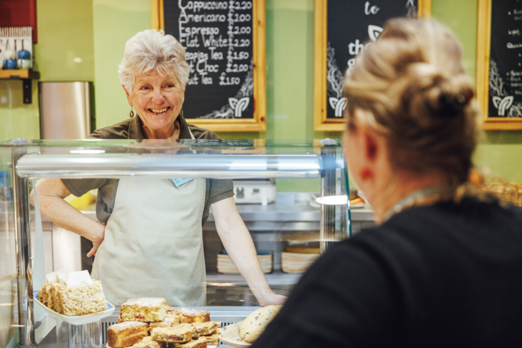 A volunteer cafe assistant wearing a green polo top and beige apron stands behind the serving counter in a cafe at St Richard's Hospice. She is serving a person and smiling a warm, friendly smile.
