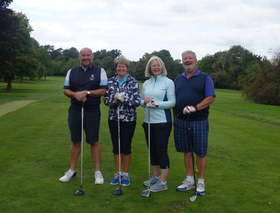 A group of four people holding golf clubs stand on the green fairway of a golf course. They are all smiling.