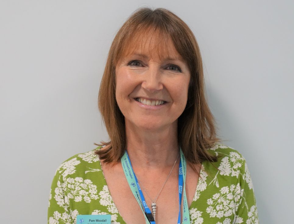 A head and shoulders photo of Pam against a plain white wall. Pam is wearing a green and white, floral printed top and is smiling.