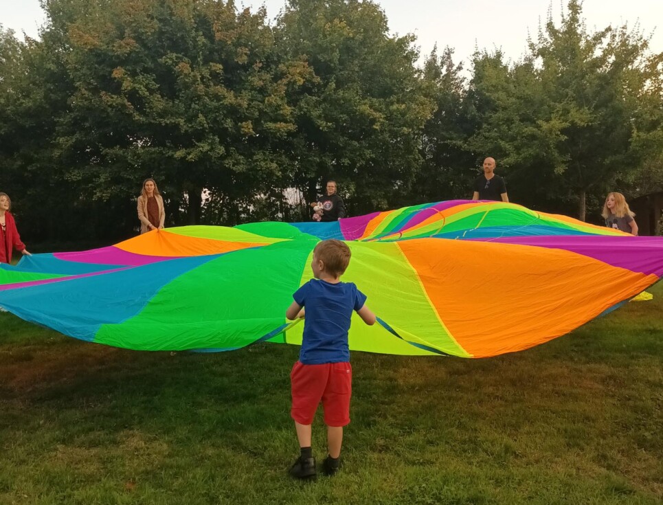 A group of children and adults attending a bereavement support group at the hospice play a game involving a large parachute in the gardens at St Richard's Hospice. They are all holding the edge of the multicoloured parachute and standing in a circle.
