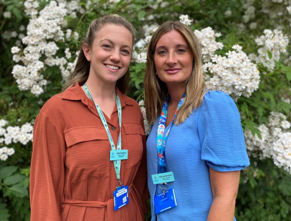 Beth and Nikki stand together by a green tree with lots of white flowers. Beth is wearing an orange dress and Nikki is wearing a bright blue top.