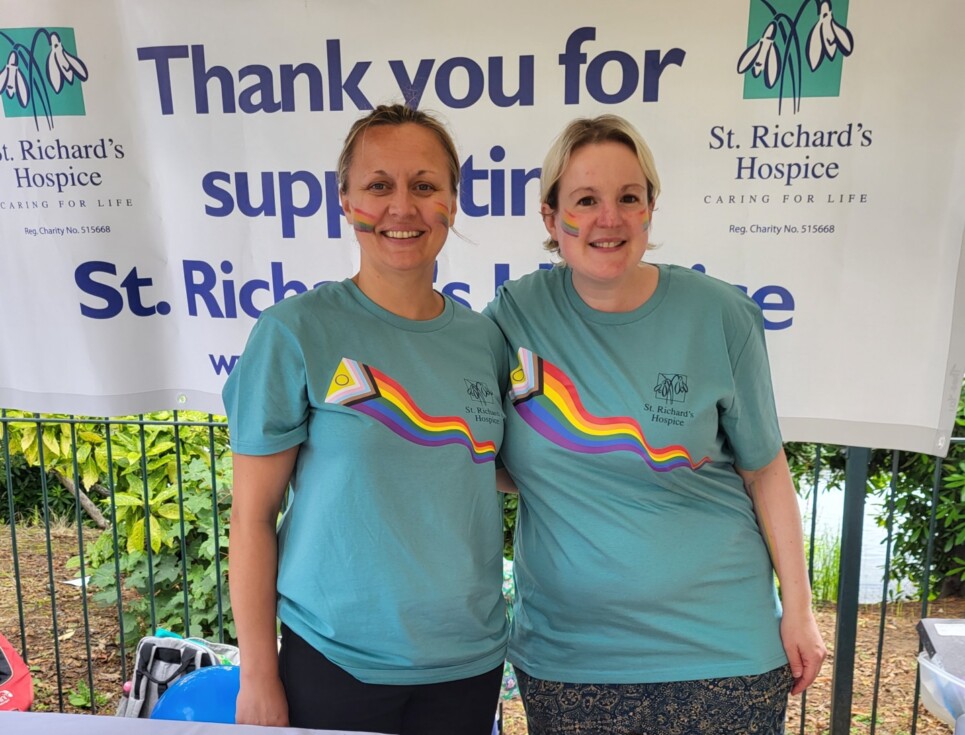 Two people wearing mint green t-shirts featuring the progress Pride flags and the St Richard's Hospice logo stand together at the St Richard's Hospice stall at Malvern Pride. Behind them is a large white banner with the text thank you for supporting St Richard's Hospice in navy writing. On the table in front of them are pictures of hospice staff at Birmingham Pride in 2019.