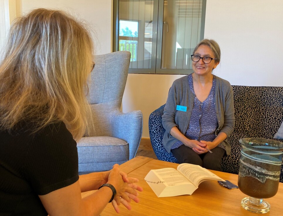 Citizens Advice Adviser Safia sits and chats to a person across a low coffee table. A large book is open on the table, next to a decorative vase. Safia is wearing a grey cardigan and purple top. The other person has their back to the camera. They are wearing a black top and have shoulder-length blonde hair.