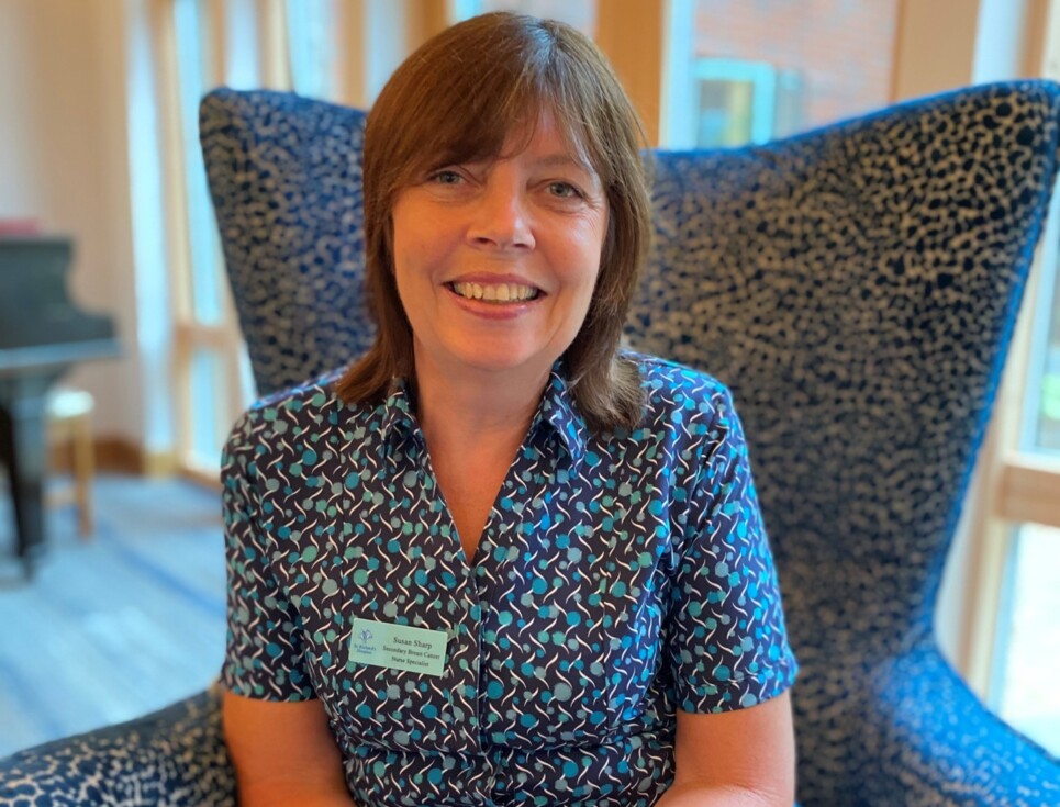 Sue sits on a comfy, high-backed armchair in the Reception area of St Richard's Hospice. Behind her are floor-to-ceiling windows. She is wearing a green and blue patterned nurses' blouse and dark trousers. The fabric of the chair is similar: a detailed dark blue and white pattern. Sue is smiling.