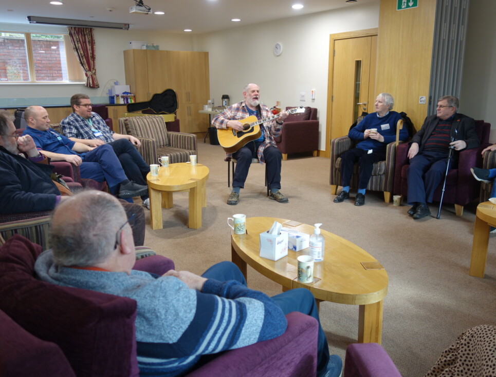 Members of the hospice's Men's Space group sit in a circle in a large room at the hospice watching musician Vo Fletcher sing and play the guitar.