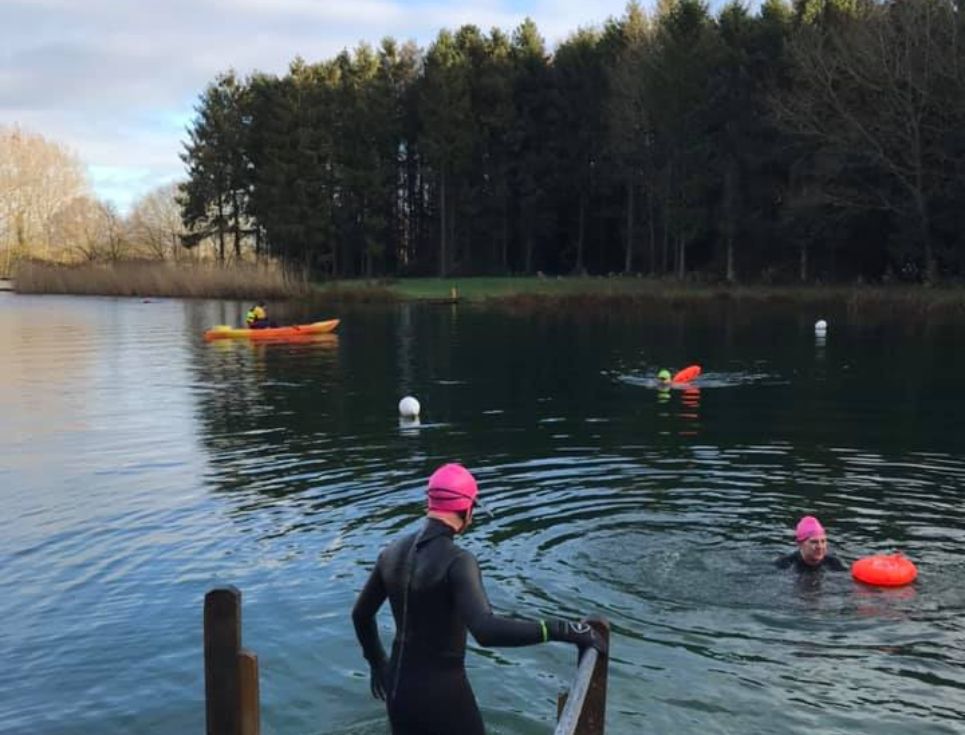 Female swimmer in a wetsuit and swim cap about to get into a lake for a 'wild swim'