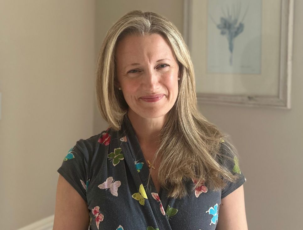 Danielle Clark pictured standing in a light sunlit room. She is wearing a grey dress decorated with images of brightly coloured butterflies. Her hair is long and blonde. Behind her is a pale coloured wall, on which hangs a picture of a bunch of flowers.
