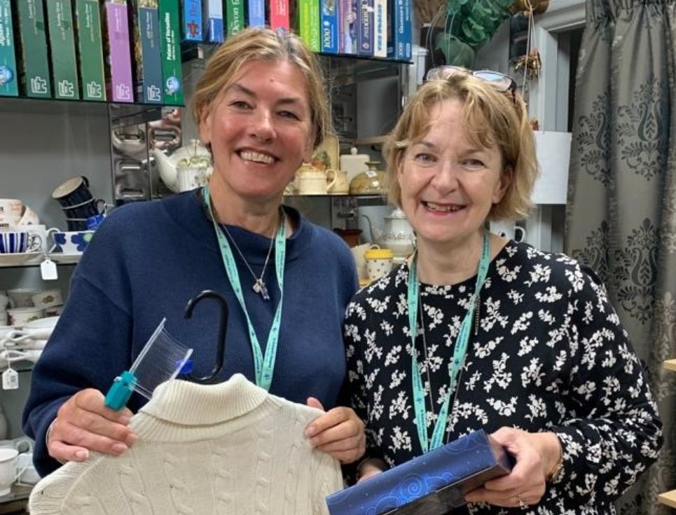 Two volunteers, Dinah and Sara, stand together holding donated items for sale in the Barnards Green shop. Behind them is a shelf of games and jigsaws. They are both smiling.