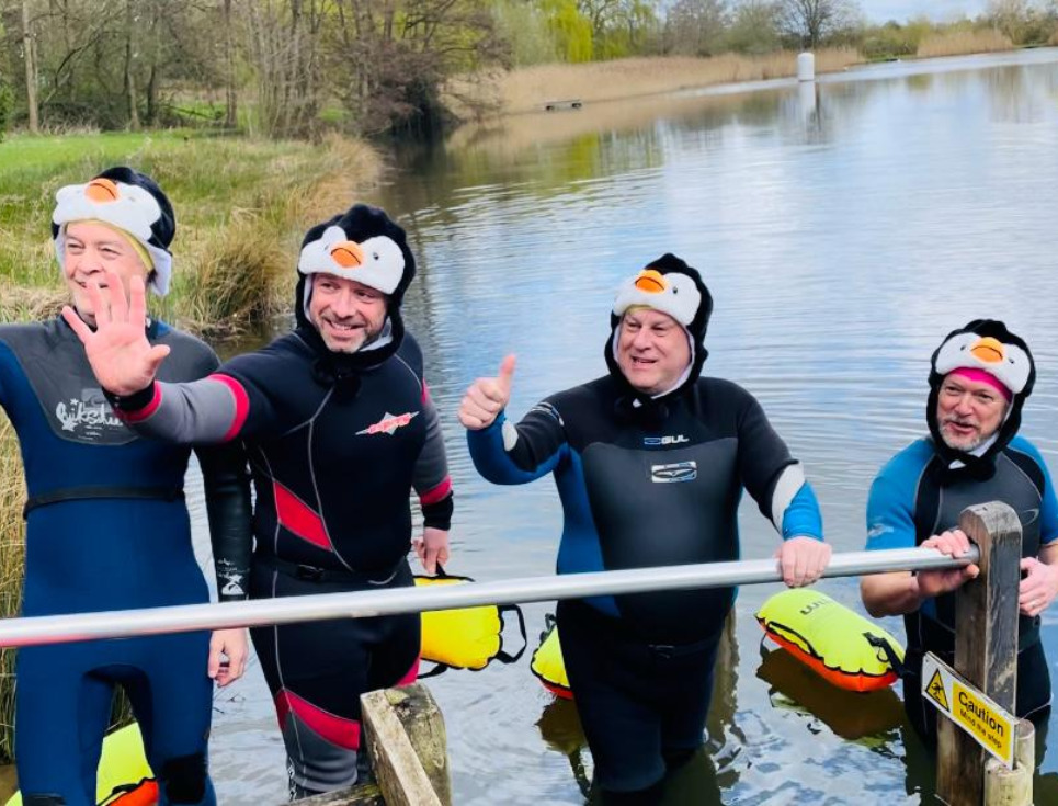 Four men in wetsuits and penguin hats submerging themselves into a fresh water lake