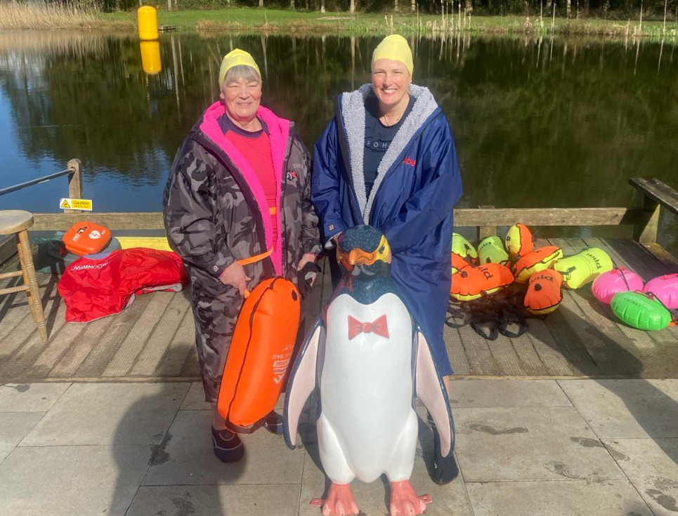 Penguin plunge two women in dry robes standing by a fresh water lake and a miniature statue of a penguin