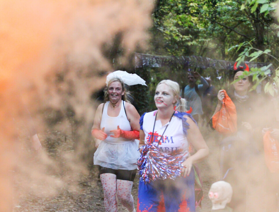 Runners take part in a previous Halloween themed run for St Richard's. Participants are dressed in Halloween outfits and are running through a cloud of orange smoke.
