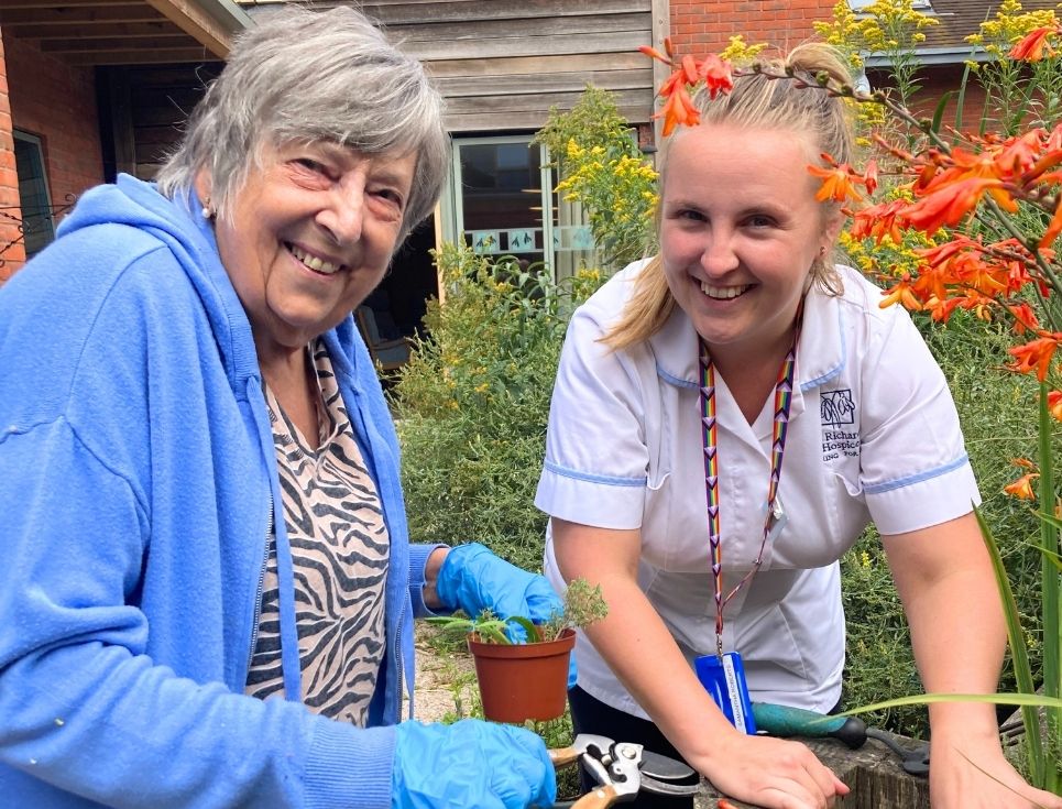 Two people, one wearing a blue hoodie and tiger print t-shirt and the other wearing a white therapy support assistant uniform, do some gardening in the courtyard gardens at St Richard's Hospice.