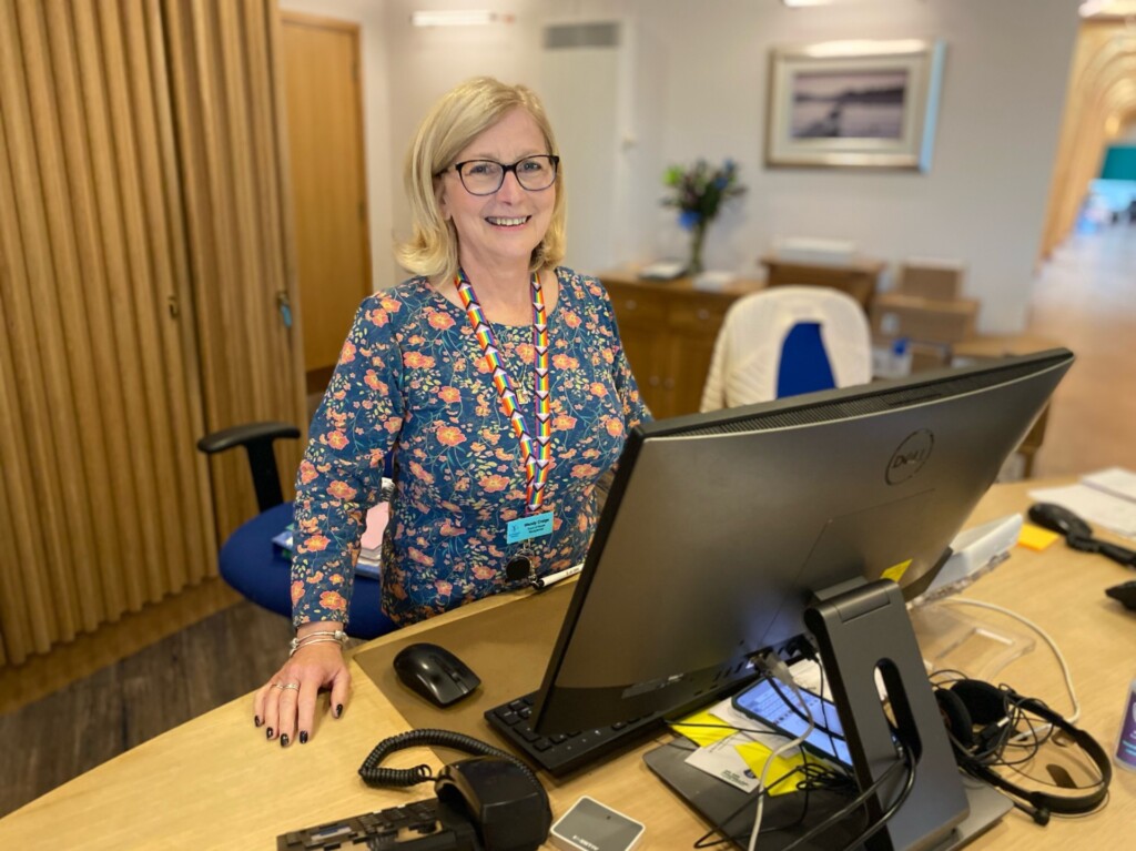 Wendy stands smiling behind the Reception desk at St Richard's Hospice. A large computer screen is on the desk in front of her, alongside a telephone. Behind her are tall wooden cabinets and the light, welcoming Reception area. Wendy has shoulder-length blonde hair and is wearing a floral top and lanyard featuring the LGBTQ+ flag colours.