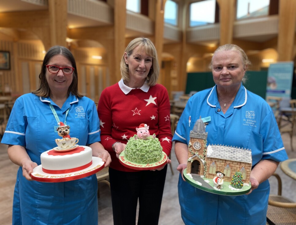 A group of three people stand together at the hospice holding intricately iced Christmas cakes. Two people are wearing blue nurses' uniforms, and the third person, standing the centre, is wearing a red jumper and black trousers. From left to right, the cakes are decorated with a pug in a tea cup, a brussels sprout with a pink pig popping out from the top, and a church.