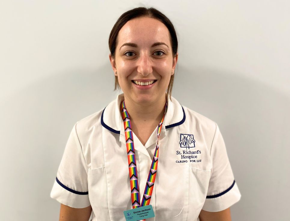 Emma pictured standing in front of a plain white wall at the hospice. She is wearing a white Physiotherapist's uniform, with dark blue edging. She also has a lanyard on in the colours of the progress Pride flag. She is smiling.