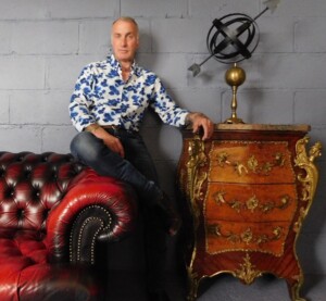 Ian Humphries pictured sitting casually on the arm of a dark red leather sofa. His arm is resting on an ornate chest of drawers which has a sculpture sitting on top. Ian is wearing a blue and white floral shirt and jeans.